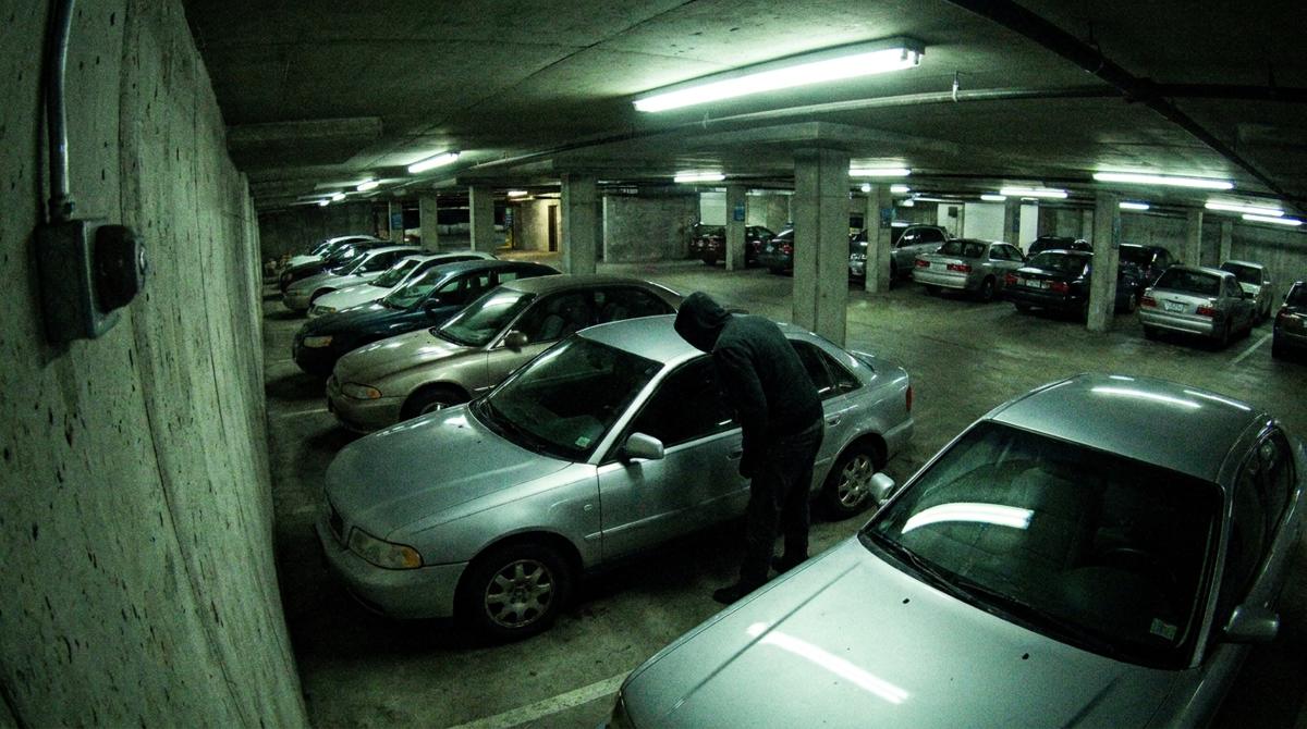 A security camera view of a person in a hooded jacket looking into a parked car’s window in a parking garage