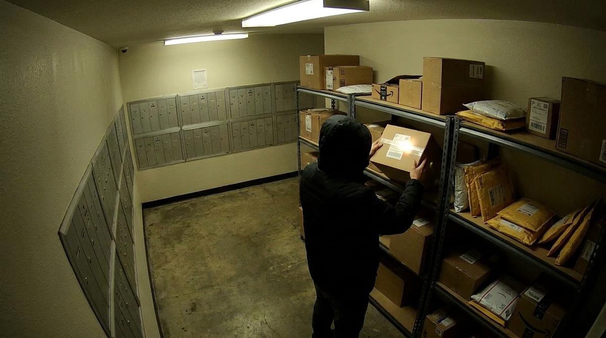 A security camera view of a person in a hooded jacket lifting a parcel from a shelf in an apartment building’s package room