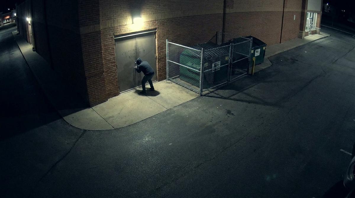 A security camera view at night of a person crouched at a store’s rear steel door beside a dumpster enclosure